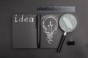 Imagem de caderno com a palavra 'Ideia' e uma ilustração de lâmpada feita com giz, além de uma lupa, uma régua e uma pasta com um clip, simbolizando o papel da IA generativa na criação de conteúdos mais ágeis e estratégicos.