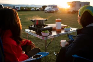 Duas pessoas desfrutando de um camping ao entardecer com um gerador portátil ligado na mesa, ideal para energia durante acampamentos.
